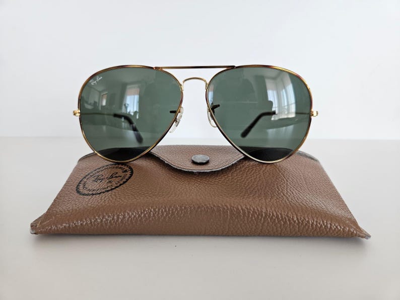 Puede incluir: Gafas de sol aviador con montura dorada y lentes verdes sobre un estuche de cuero marr&oacute;n. El estuche tiene un cierre de bot&oacute;n a presi&oacute;n y un logotipo circular. Las gafas reflejan el entorno.