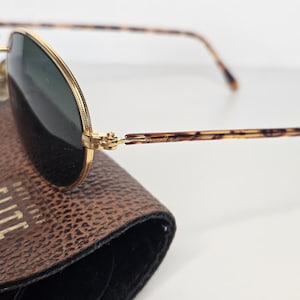 May include: Gold-framed sunglasses with dark green lenses and tortoiseshell temples. The sunglasses are on a brown leather case. The case has the word "ELITE" printed on it.