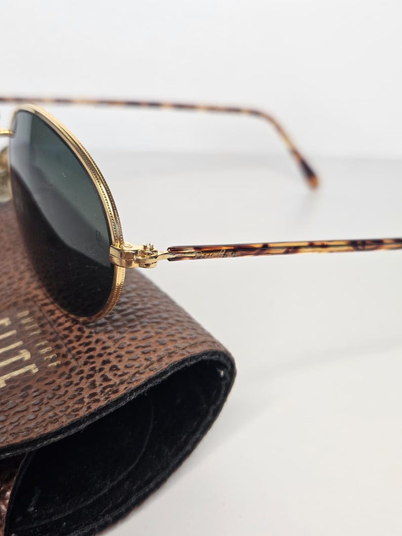 May include: Gold-framed sunglasses with dark green lenses and tortoiseshell temples. The sunglasses are on a brown leather case. The case has the word "ELITE" printed on it.