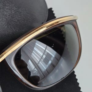 Pu&ograve; includere: Occhiali da sole con montatura tartarugata marrone scuro e un dettaglio in metallo dorato lucido sulla parte superiore. Le lenti sono scure, colorate e riflettenti, mostrando un leggero riflesso dell'ambiente circostante. Poggiano su un panno nero.