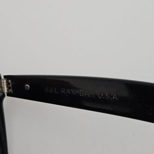 May include: Close-up of a black Ray-Ban sunglasses arm. The text "B&L RAY-BAN U.S.A." is printed in white on the arm. The hinge and a small silver rivet are visible.