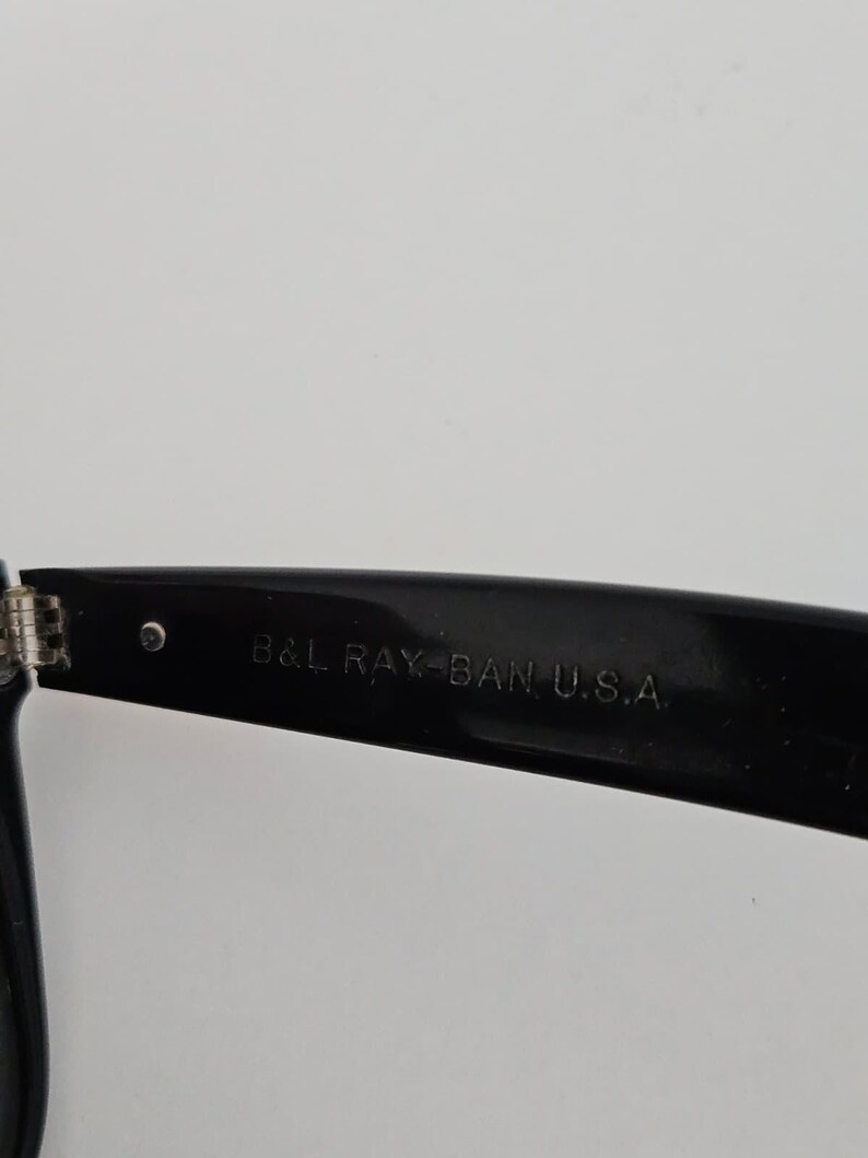 May include: Close-up of a black Ray-Ban sunglasses arm. The text "B&L RAY-BAN U.S.A." is printed in white on the arm. The hinge and a small silver rivet are visible.