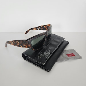 May include: Tortoiseshell sunglasses with dark lenses rest on a black leather case. A gray cleaning cloth with a red logo is next to the case. The sunglasses have a rectangular frame and a classic design.