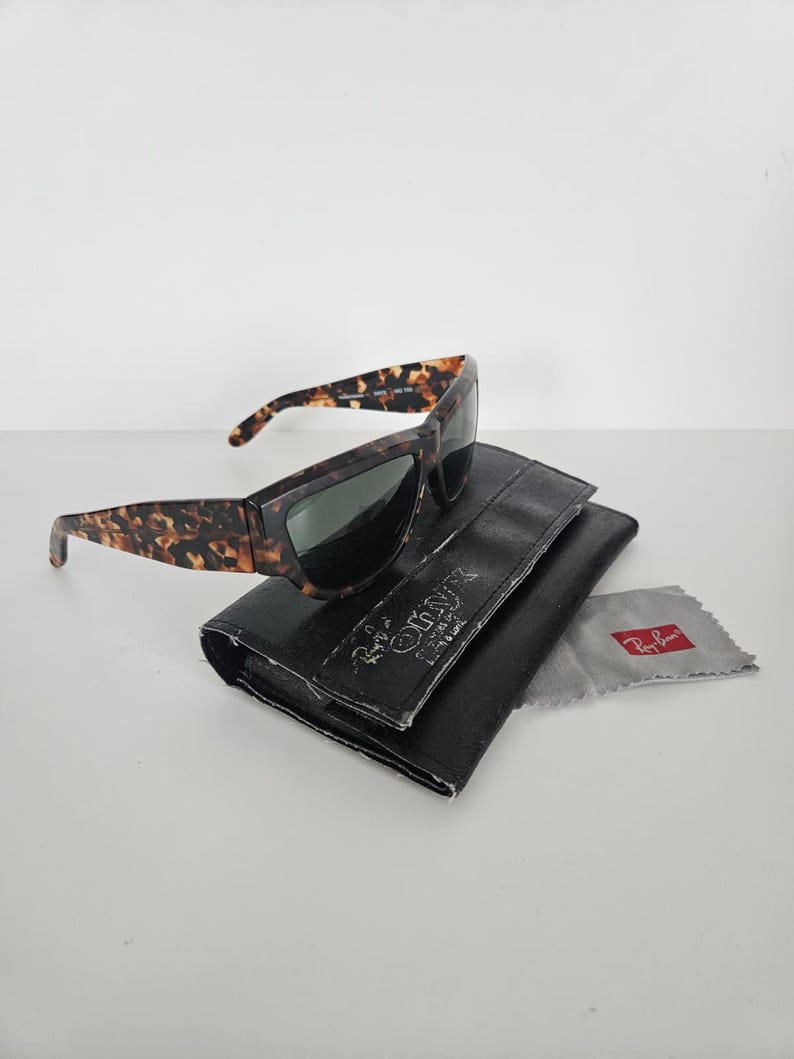 May include: Tortoiseshell sunglasses with dark lenses rest on a black leather case. A gray cleaning cloth with a red logo is next to the case. The sunglasses have a rectangular frame and a classic design.