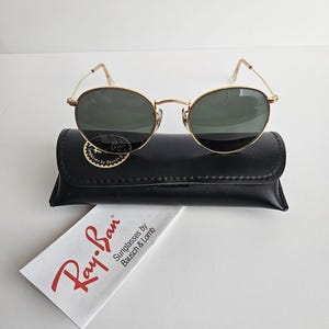 May include: Gold-framed Ray-Ban sunglasses with dark lenses rest on a black leather case. A white cleaning cloth with the Ray-Ban logo and text is visible. The sunglasses have a classic, oval shape.