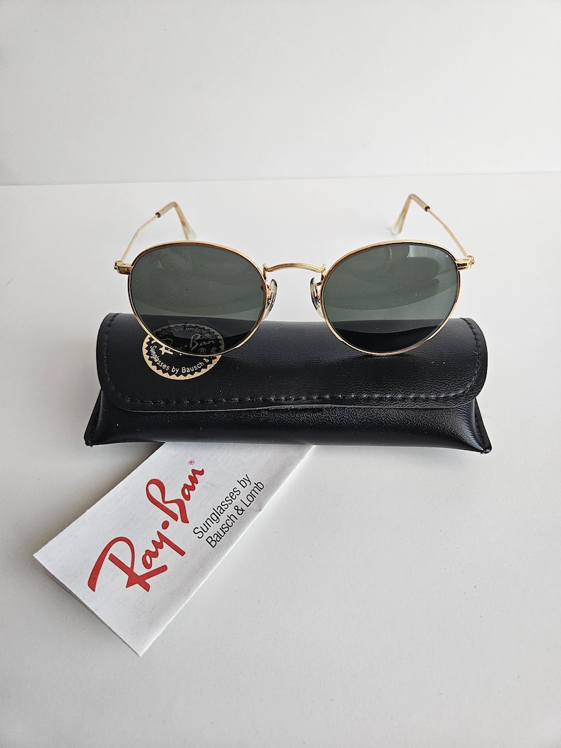 May include: Gold-framed Ray-Ban sunglasses with dark lenses rest on a black leather case. A white cleaning cloth with the Ray-Ban logo and text is visible. The sunglasses have a classic, oval shape.