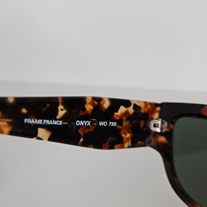 May include: Close-up of sunglasses with a tortoiseshell pattern on the frame. The text "FRAME FRANCE - ONYX WO 796" is printed on the arm. The lenses are dark green.