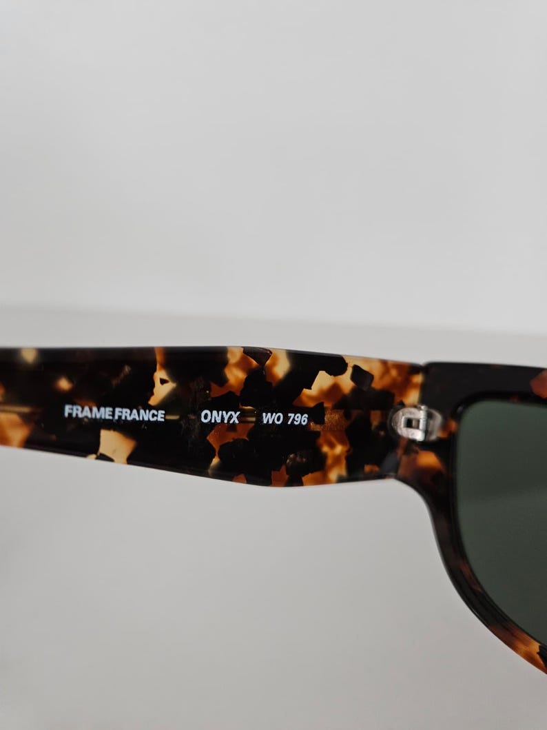 May include: Close-up of sunglasses with a tortoiseshell pattern on the frame. The text "FRAME FRANCE - ONYX WO 796" is printed on the arm. The lenses are dark green.