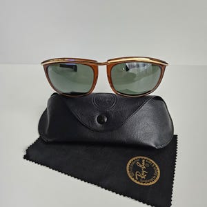 Pu&ograve; includere: Occhiali da sole Ray-Ban con montatura color ambra, barra frontale in metallo dorato e lenti verde scuro. Gli occhiali sono posizionati sopra una custodia in similpelle nera e un panno in microfibra nero con un logo dorato.