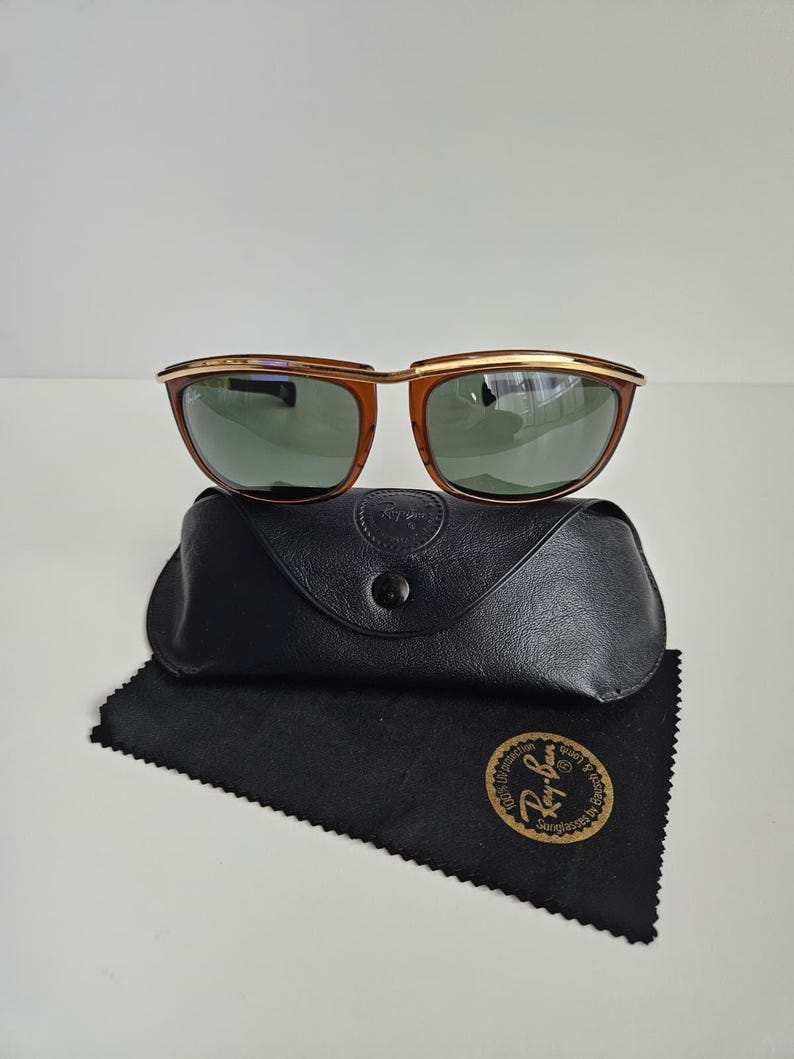 Pu&ograve; includere: Occhiali da sole Ray-Ban con montatura color ambra, barra frontale in metallo dorato e lenti verde scuro. Gli occhiali sono posizionati sopra una custodia in similpelle nera e un panno in microfibra nero con un logo dorato.