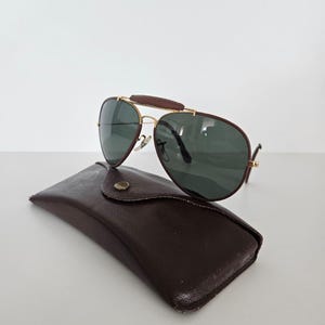Puede incluir: Gafas de sol estilo aviador con montura dorada, lentes verde oscuro y puente de cuero marr&oacute;n. Descansan sobre un estuche de cuero marr&oacute;n con cierre de bot&oacute;n. Dise&ntilde;o cl&aacute;sico y elegante.