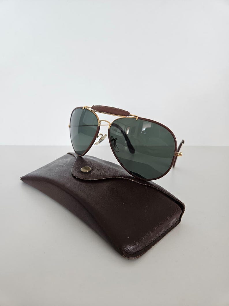Puede incluir: Gafas de sol estilo aviador con montura dorada, lentes verde oscuro y puente de cuero marr&oacute;n. Descansan sobre un estuche de cuero marr&oacute;n con cierre de bot&oacute;n. Dise&ntilde;o cl&aacute;sico y elegante.