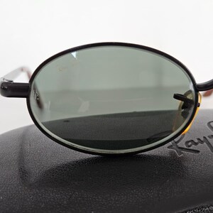 Puede incluir: Gafas de sol ovaladas con montura negra y lentes verdes. Las gafas de sol est&aacute;n sobre un estuche negro. El logotipo de Ray-Ban es visible en el estuche.