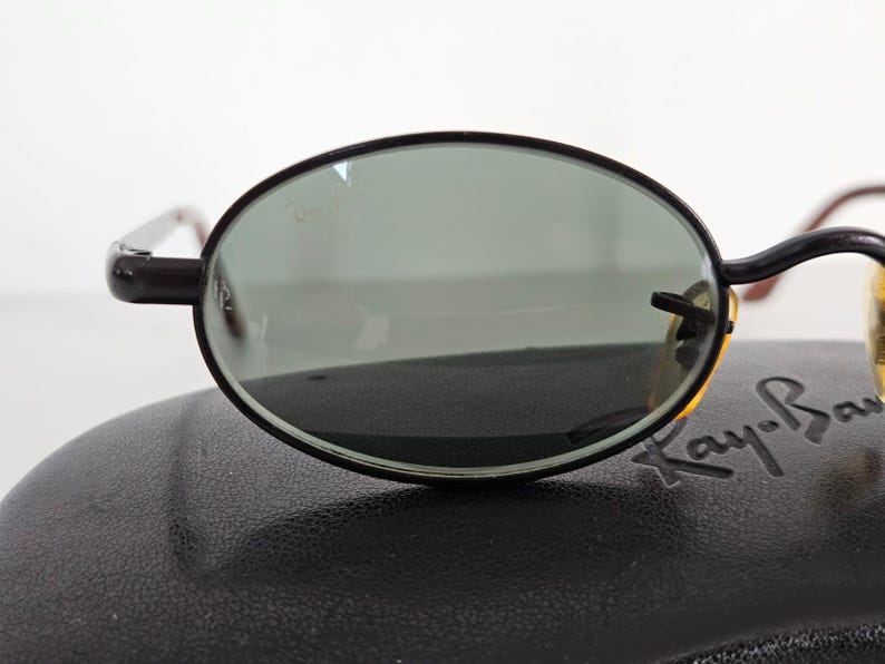 Puede incluir: Gafas de sol ovaladas con montura negra y lentes verdes. Las gafas de sol est&aacute;n sobre un estuche negro. El logotipo de Ray-Ban es visible en el estuche.
