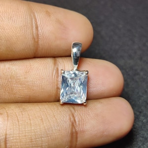 May include: A rectangular, clear gemstone pendant with a silver bail. The gemstone is set in a silver frame and has a brilliant cut. The pendant is held in a hand.