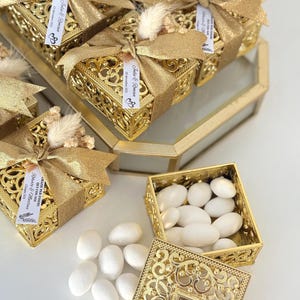 Pode incluir: Caixas douradas ornamentadas com tampas, amarradas com fita dourada e adornadas com flores secas. As caixas contêm doces brancos. Algumas caixas têm etiquetas com texto.