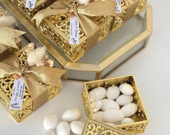 May include: Gold-colored, ornate boxes with lids, tied with gold ribbon and adorned with dried flowers. The boxes are filled with white candies. Some boxes have tags with text.