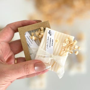 May include: Small, rectangular soaps wrapped with cream-colored ribbon and dried flowers. Each soap has a small tag that reads "Ana & Oscar Our Wedding 02.04.2025". The soaps are presented in a small, brown cardboard box.