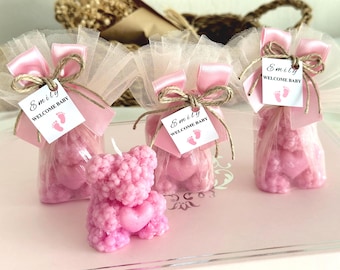 May include: Pink baby shower favors. The image shows a pink teddy bear candle with a heart and three small bags of pink candies. Each bag is tied with a pink ribbon and a tag that says "Emily Welcome Baby" with baby footprints.