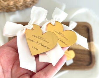 May include: Gold heart-shaped tags with white ribbon bows. The tags have the text "THANK YOU FOR CELEBRATING WITH US" and names and a date. The tags are on a wooden tray.