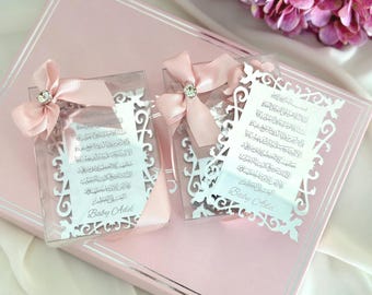 May include: Two clear acrylic favors with intricate white cut-out designs and Arabic script, tied with pink ribbons and crystal accents. The favors are presented on a pink box, with a floral arrangement in the background. The text "Baby Adel" is visible.