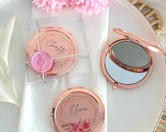 Peut inclure: Miroirs compacts rose gold avec des motifs floraux et des noms personnalisés, présentés sur une assiette blanche avec des décorations florales roses. Un miroir est ouvert, révélant une surface réfléchissante. Un autre est emballé dans une boîte transparente avec un sceau de cire.