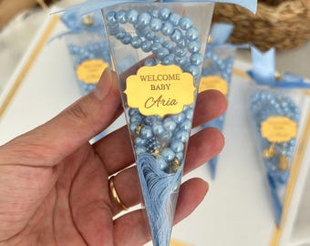 May include: A clear, cone-shaped favor box tied with a light blue ribbon and a decorative jewel. Inside, a light blue beaded rosary is visible. The box has a gold label that reads "WELCOME BABY Aria".