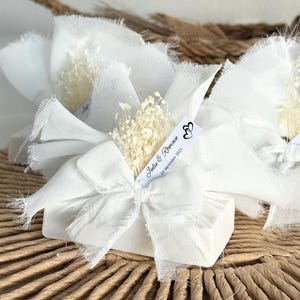 Puede incluir: Cajas de regalo blancas envueltas en tela blanca y atadas con lazos. Cada caja está adornada con flores secas y una etiqueta que dice "Julie & Romain 20 décembre 2025". Las cajas están dispuestas sobre una superficie tejida.