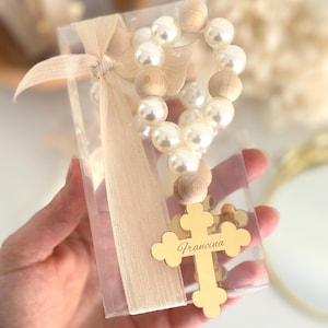 Peut inclure: Un chapelet avec des perles blanches et des perles en bois, présenté dans une boîte transparente nouée avec un ruban beige. Une croix dorée avec le nom "Francina" est attachée au chapelet.