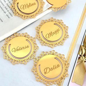 May include: Gold-colored metal bookmarks with intricate, circular lace-like edges. Each bookmark features a name engraved in a cursive font: Melina, Maggi, Valeria, Dalia, and Martine. The bookmarks are arranged on a book and a white surface.