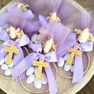 Puede incluir: Primer plano de recuerdos de bautizo. Cada recuerdo incluye una cruz dorada con el nombre "Giuliana Grace" y la fecha "9-21-25". Los recuerdos están decorados con cintas lavanda, tul y flores secas, dispuestos en una bandeja.