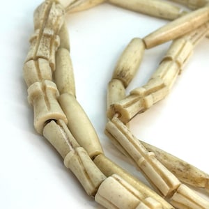 May include: A necklace composed of off-white, cylindrical beads with a carved, ribbed texture. The beads are strung together, displaying a weathered, organic appearance with subtle brown flecks and variations in the material surface.