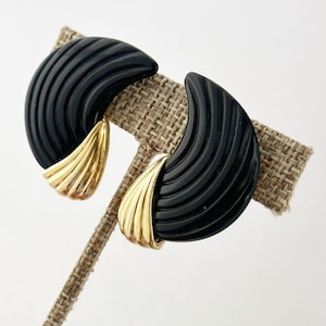 May include: A pair of black, half-moon shaped earrings with a ridged texture. The earrings have a gold-colored accent on the lower portion. The earrings are displayed on a tan, textured surface.