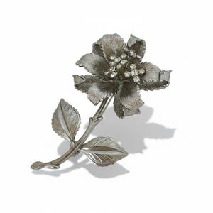 May include: A silver-toned floral brooch with a flower and two leaves. The flower has multiple petals and is adorned with small, clear crystals at its center. The leaves and stem are detailed with textured lines.