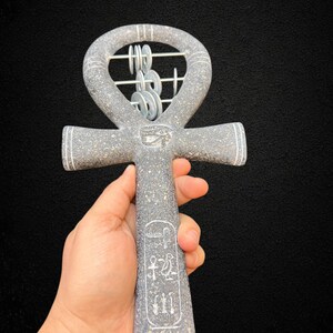 May include: A gray stone ankh with white accents and hieroglyphs. The ankh features a loop at the top with a decorative silver element. The ankh is held against a black background.