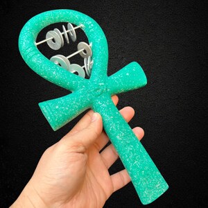 May include: A turquoise-colored ankh-shaped musical instrument with silver metal discs. The ankh is held in a hand against a black background. The instrument has a textured surface.
