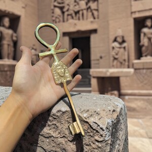 May include: A gold-colored Ankh key, held in a hand, with a detailed pharaoh face on the key's head. The key is set against a backdrop of an ancient stone structure, suggesting an Egyptian theme.