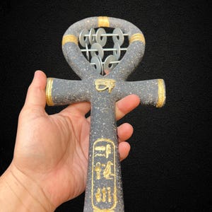 Pode incluir: Um objeto decorativo cinza em forma de ankh com detalhes dourados. O ankh apresenta um laço na parte superior com pequenos discos prateados. Detalhes dourados estão presentes nos braços da cruz e no laço superior. Hieróglifos são visíveis.