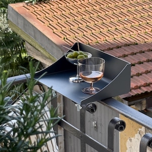 K&ouml;nnte beinhalten: Ein dunkelgraues Balkontablett h&auml;lt ein Glas Ros&eacute;wein und eine kleine Schale mit gr&uuml;nen Oliven. Das Tablett ist an einem schmiedeeisernen Gel&auml;nder befestigt, mit einem roten Ziegeldach im Hintergrund. Ein Rosmarinzweig befindet sich im Vordergrund.