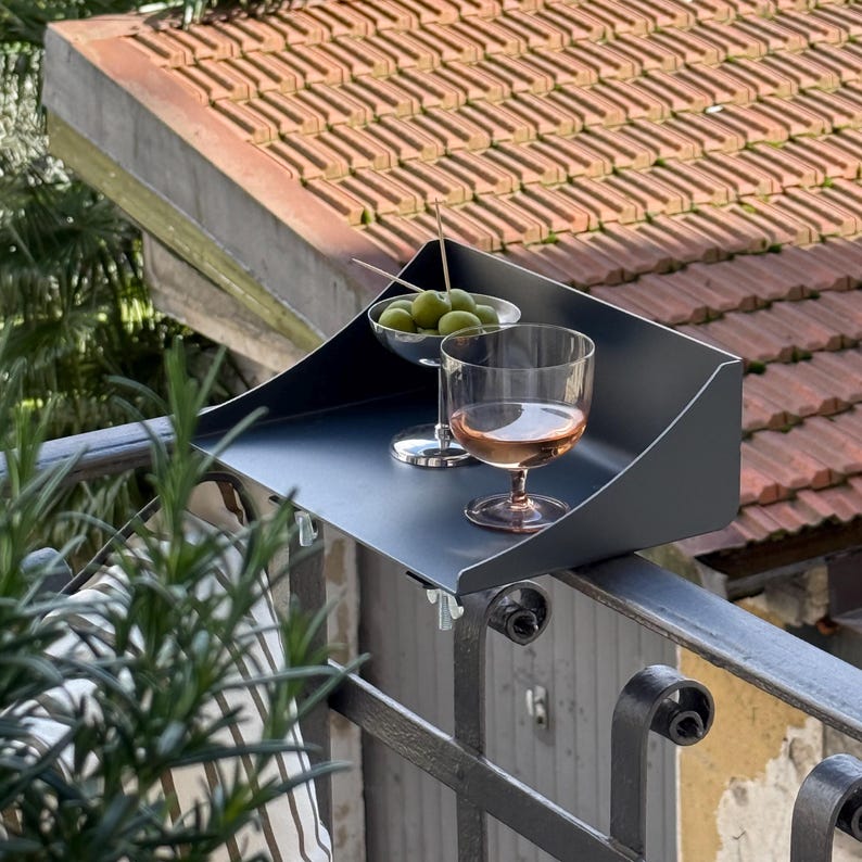 K&ouml;nnte beinhalten: Ein dunkelgraues Balkontablett h&auml;lt ein Glas Ros&eacute;wein und eine kleine Schale mit gr&uuml;nen Oliven. Das Tablett ist an einem schmiedeeisernen Gel&auml;nder befestigt, mit einem roten Ziegeldach im Hintergrund. Ein Rosmarinzweig befindet sich im Vordergrund.