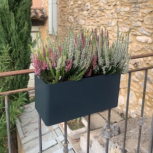 Puede incluir: Una jardinera rectangular gris oscuro llena de vibrantes plantas de brezo. El brezo presenta flores rosas y blancas, que contrastan con el recipiente oscuro. La jardinera está montada en una barandilla de hierro forjado, con una pared de piedra al fondo.