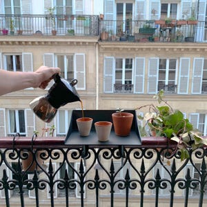 K&ouml;nnte beinhalten: Eine schwarze und silberne Espressokanne gie&szlig;t Kaffee in zwei kleine Keramikbecher auf ein schwarzes Tablett. Ein Terrakottatopf mit einer kleinen Pflanze steht auf dem Tablett, mit einem Balkon und einem Geb&auml;ude im Hintergrund.
