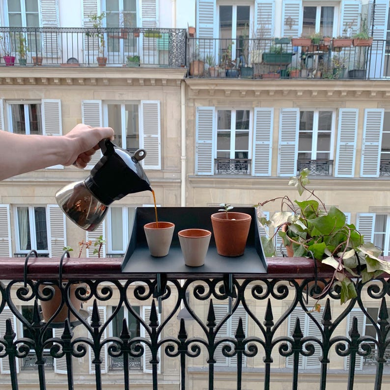 K&ouml;nnte beinhalten: Eine schwarze und silberne Espressokanne gie&szlig;t Kaffee in zwei kleine Keramikbecher auf ein schwarzes Tablett. Ein Terrakottatopf mit einer kleinen Pflanze steht auf dem Tablett, mit einem Balkon und einem Geb&auml;ude im Hintergrund.