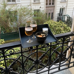 May include: A black balcony tray holds two glasses of amber liquid, a small bowl of snacks, and a candle. The tray is attached to a black wrought iron railing, with a background of greenery and a building.
