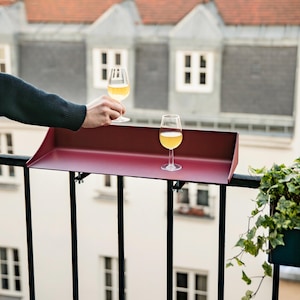 Könnte beinhalten: Ein dunkelrotes Balkonregal aus Metall mit rechteckiger Ablagefläche und erhöhtem hinteren Rand, das sicher an schwarzen vertikalen Geländerstäben befestigt ist. Auf dem Regal steht ein klares Glas mit einer hellgelben Flüssigkeit.