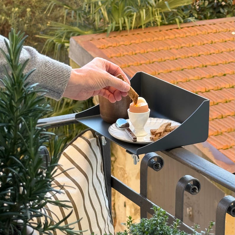 K&ouml;nnte beinhalten: Ein schwarzes Metall-Balkontablett h&auml;lt ein weich gekochtes Ei in einem wei&szlig;en Eierbecher, Toast und einen kleinen L&ouml;ffel. Das Tablett ist an einem schwarzen Metallgel&auml;nder befestigt. Eine Person taucht Toast in das Ei. Der Hintergrund umfasst ein rotes Ziegeldach und Gr&uuml;n.