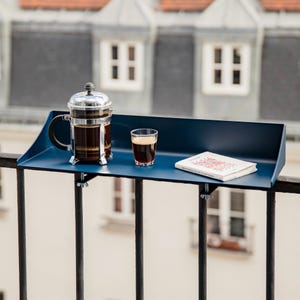 Könnte beinhalten: Ein dunkelblaues Balkonregal aus Metall mit rechteckiger Ablagefläche und erhöhtem hinteren Rand. Das Regal ist mit zwei silbernen Flügelmuttern an einem schwarzen Metallgeländer befestigt. Eine French Press, ein Glas Kaffee und ein Buch stehen darauf.