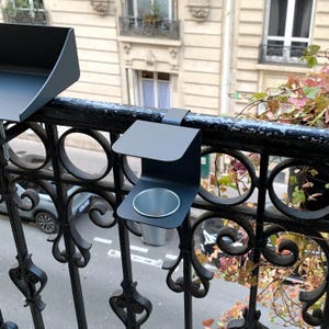 Könnte beinhalten: Schwarzer Metall-Balkon-Becherhalter mit kreisförmiger Basis und gebogener Oberseite, befestigt an einem schwarzen schmiedeeisernen Geländer. Der Halter ist zum Aufhängen über dem Geländer konzipiert. Der Hintergrund zeigt ein Gebäude mit Fenstern und einer Straße.