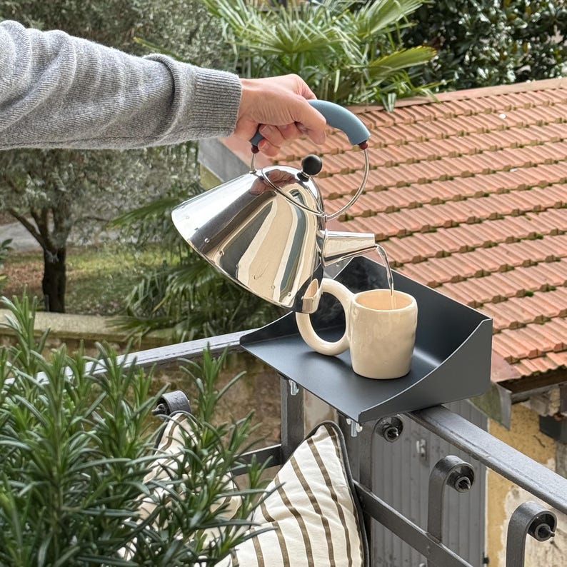 K&ouml;nnte beinhalten: Ein Edelstahl-Teekessel mit blauem Griff gie&szlig;t Wasser in eine cremefarbene Tasse. Die Tasse steht auf einem schwarzen Tablett, das an einem Metallgel&auml;nder befestigt ist. Der Hintergrund zeigt Gr&uuml;n und ein rotes Ziegeldach.