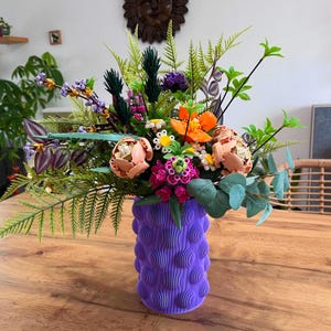 Peut inclure: Un bouquet de fleurs artificielles colorées dans un vase violet texturé. L'arrangement comprend des fleurs pêche, orange, roses et blanches, avec un feuillage vert. Le vase a un design arrondi unique. Le bouquet est sur une table en bois.